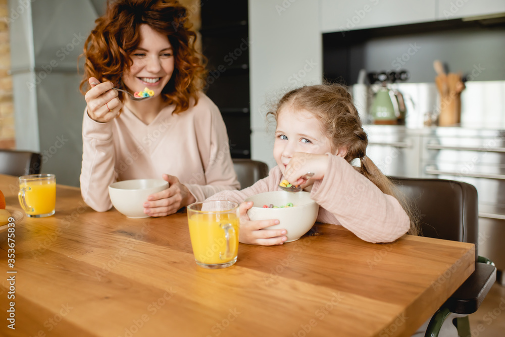 cheerful mother and daughter holding spoons near bowls with corn flakes and glasses of orange juice cheerful mother and daughter holding spoons near bowls with corn flakes and glasses of orange juice