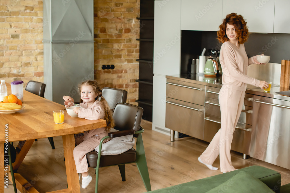 happy kid near bowl with corn flakes, glass of orange juice and curly mother in kitchen happy kid near bowl with corn flakes, glass of orange juice and curly mother in kitchen