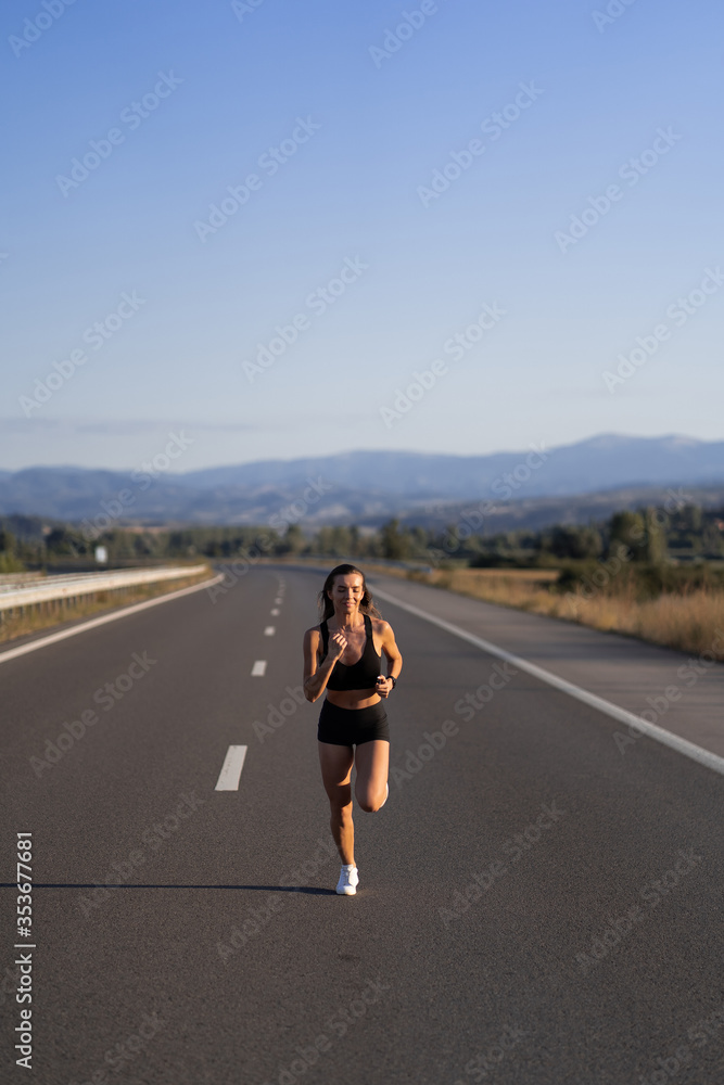 Young girl running along the road. Athletic happy woman jogging in trendy black sexy top and shorts enjoying the sun exercising. Healthy lifestyle. Perfect fitness body shapes and tan skin