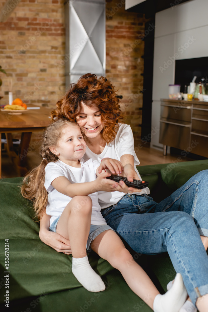 cute kid holding remote controller and sitting near cheerful mother on sofa cute kid holding remote controller and sitting near cheerful mother on sofa