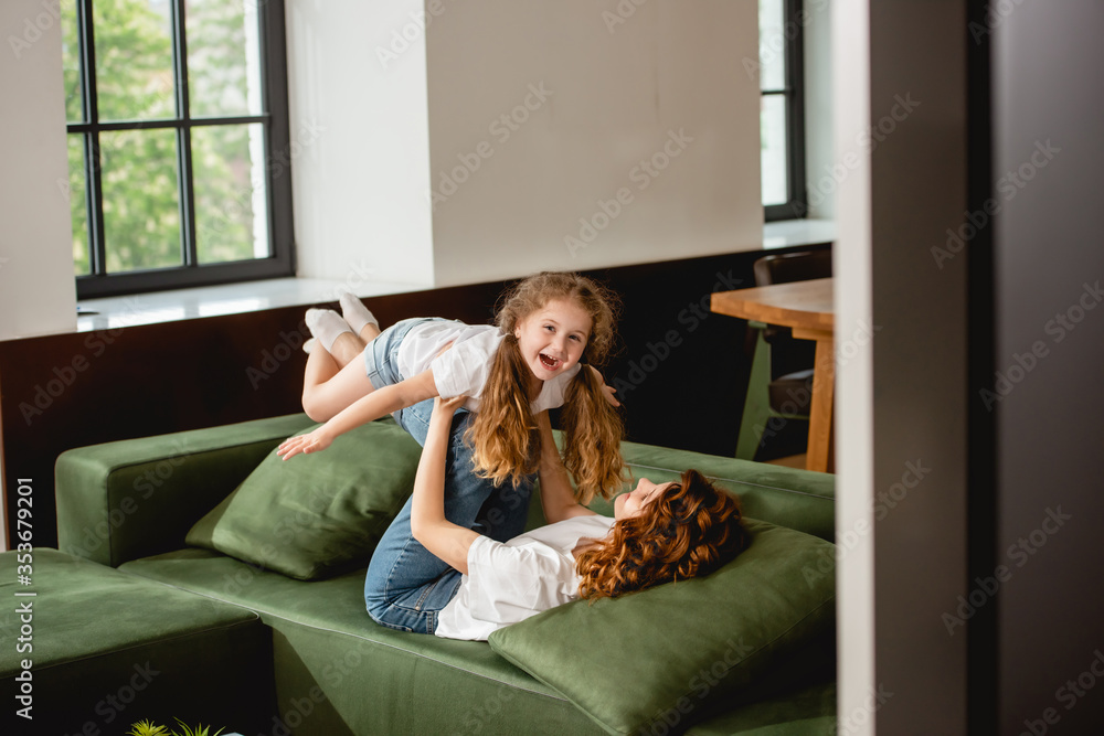 selective focus of curly mother lying on sofa, lifting happy daughter and holding hands in living room selective focus of curly mother lying on sofa, lifting happy daughter and holding hands in living room