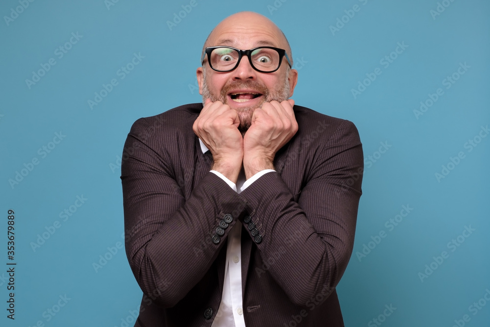 Handsome mature man looks with excitement holding fists near face,receiving a great news, isolated over blue wall. Studio shot on blue wall. Handsome mature man looks with excitement holding fists near face,receiving a great news, isolated over blue wall. Studio shot on blue wall.