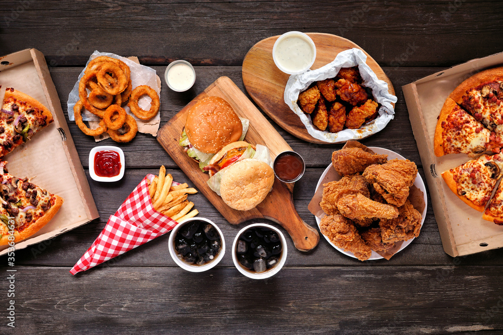 Table scene with large variety of take out and fast foods. Hamburgers, pizza, fried chicken and sides. Above view on a dark wood background. - obrazy, fototapety, plakaty 