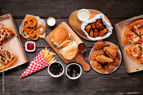 Table scene with large variety of take out and fast foods. Hamburgers, pizza, fried chicken and sides. Above view on a dark wood background.