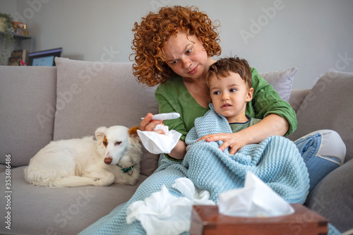 Photo of worried mother taking temperature of her little son who is lying in bed with fever. Mother checking the temperature of her ill boy with thermometer on a couch in the living room at home.
