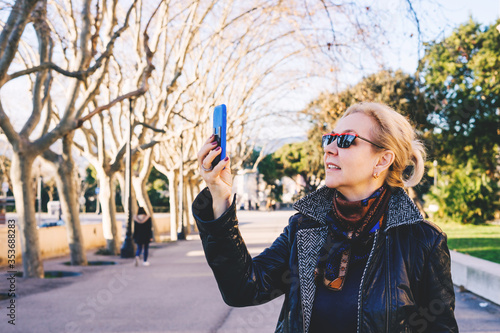 An adult blonde lady use Facetime to talking on the phone in a dark coat and glasses looks up in spring park and smiles. Side view.