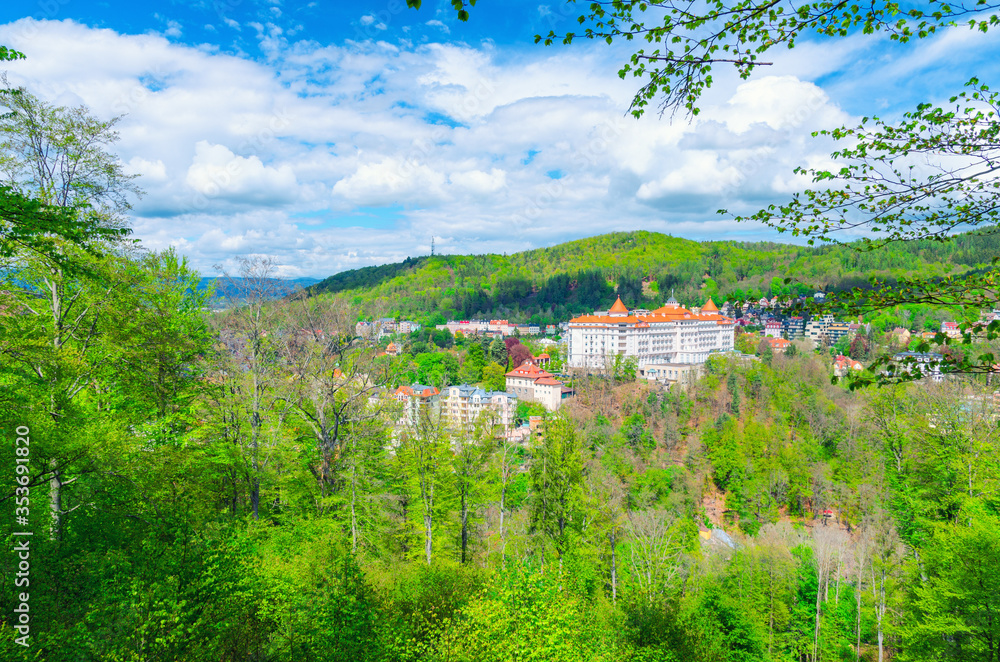 Karlovy Vary (Carlsbad) historical city centre top aerial view with hotels buildings, Slavkov Forest hills with green trees on slope, blue sky white clouds background, West Bohemia, Czech Republic