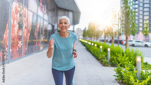 Front View Of Senior Woman Jogging Through Park. Elderly woman running with smartwatches in the park in evening sunset. Attractive looking mature woman keeping fit and healthy. © Dragana Gordic