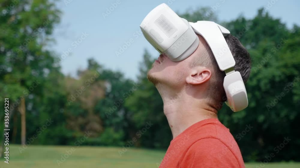 Man in a virtual reality helmet raises his head up. Guy in a park in a red T-shirt on background of green trees looks VR glasses. 4K footage Man in a virtual reality helmet raises his head up. Guy in a park in a red T-shirt on background of green trees looks VR glasses. 4K footage