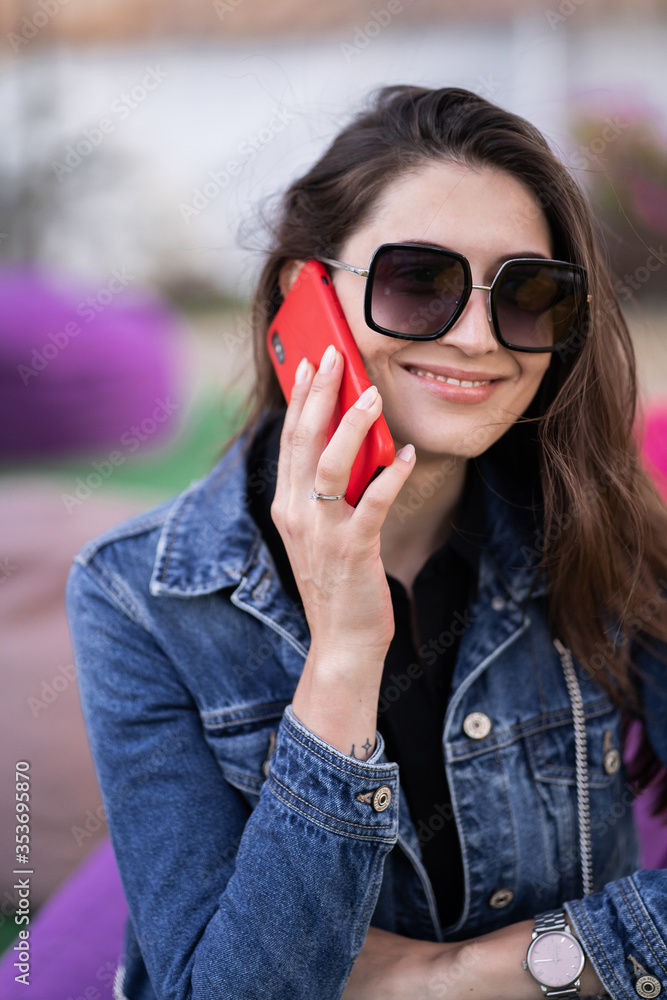 Attractive girl resting on nature at weekend. Girl in jeans jacket sitting in park. Caucasian brunette young woman talking on smartphone outside alone.Technologies and gadgets. Attractive girl resting on nature at weekend. Girl in jeans jacket sitting in park. Caucasian brunette young woman talking on smartphone outside alone.Technologies and gadgets.