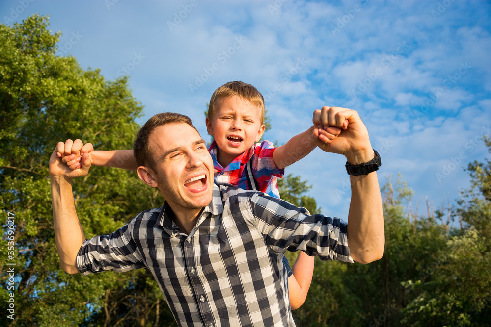 Happy young father holds his son piggyback ride on his shoulders and looking up. Little boy is sitting piggyback on shoulders his dad