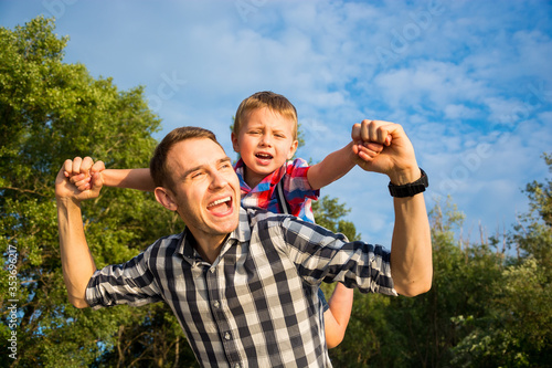 Obraz na płótnie Happy young father holds his son piggyback ride on his shoulders and looking up. Little boy is sitting piggyback on shoulders his dad w salonie