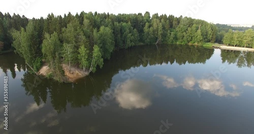 4K aerial summer early morning high quality video footage of pristine lush green pine tree surrounded forest lake with dark calm water reflecting clouds, a dam, island and a sand beach near small town