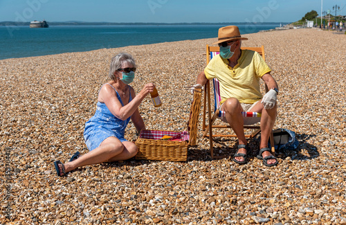 Southsea, Portsmouth, Southern England, UK. May 2020. Woman  wearing a surgical mask  social distancing from her husband during the Corvid-19 outbreak. Snacking on the beach in Southsea, UK. photo