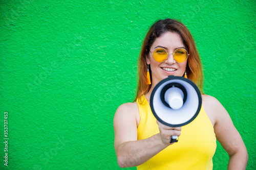 Happy woman in a yellow dress glasses and earrings on a green background shouting into a megaphone. Portrait of a girl holding a loudspeaker. Lady with a perfect snow-white smile.