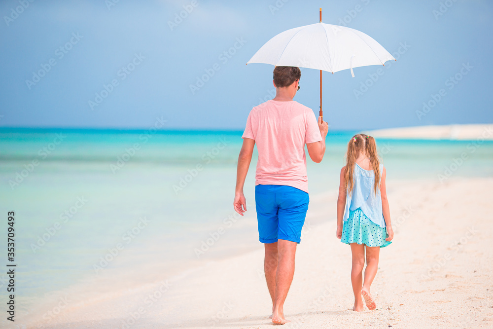 Young father and his little daughter walking under umbrella on white sand beach