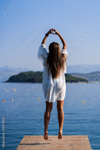 Young beautiful woman in white dress jumping on pier with seaview background and showing heart from fingers. The concept of joy, ease and freedom during the vacation. The girl is enjoying the rest