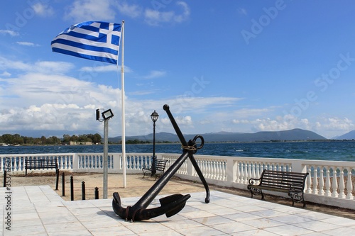 Greek flag behind a large old anchor at the sailors monument town square, in Preveza, Greece. View of Aktio and Ambracian gulf at the background