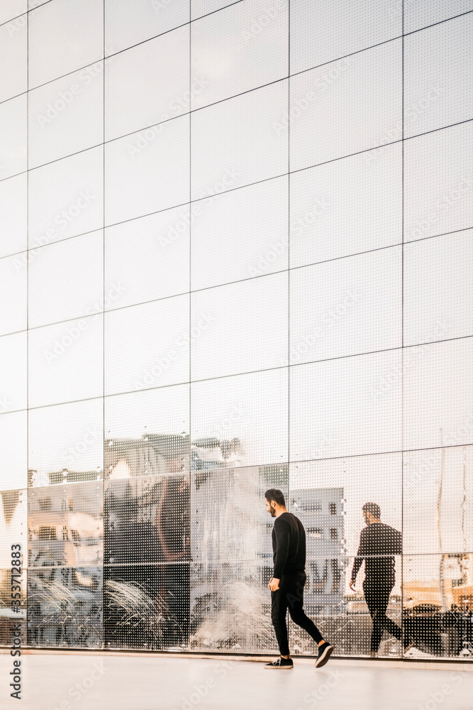 Full length side view of young man walking by reflection on glass building Full length side view of young man walking by reflection on glass building