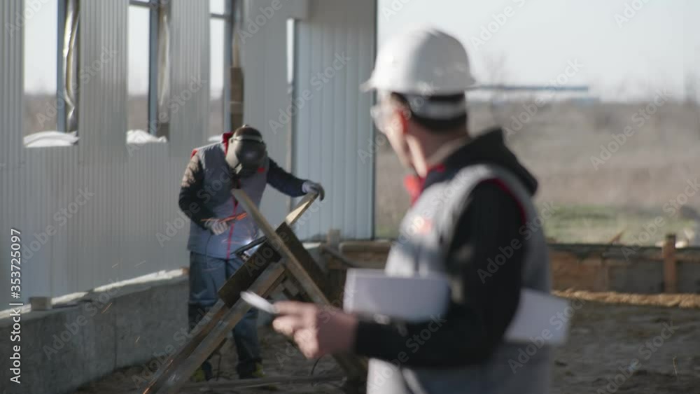 portrait of smiling male engineer in protective helmet with drawings in his hands, smiles and inspects construction of new hangar background of a welder portrait of smiling male engineer in protective helmet with drawings in his hands, smiles and inspects construction of new hangar background of a welder