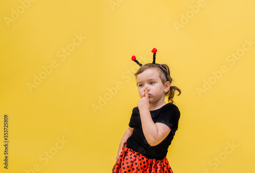 A pensive little girl in a Ladybird costume points her finger "Quietly" against a yellow isolated background with space for text.
