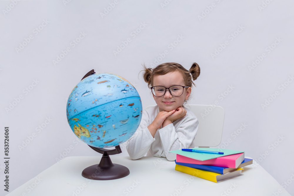Portrait of a small pupil with glasses sitting at a table on a white isolated background with space for text. On the table is a globe, books and a notebook with a pen. Portrait of a small pupil with glasses sitting at a table on a white isolated background with space for text. On the table is a globe, books and a notebook with a pen.