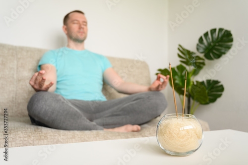 A man with closed eyes sits on a sofa in the lotus position and meditates at home. Smoldering incense stick on the table. Selective focus.