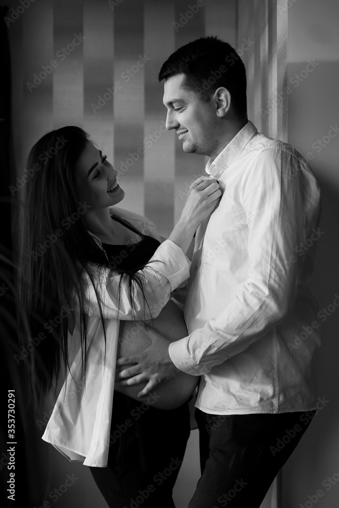 Romantic couple expecting baby, holding hands and touching foreheads while standing together against window at home, free space. Pregnancy, maternity, preparation and expectation concept. C