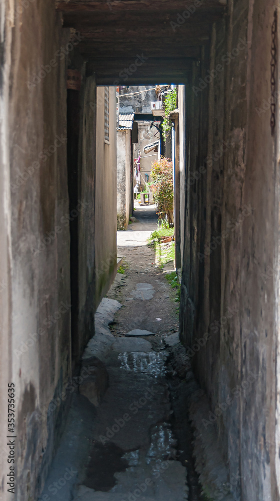 Obraz premium Tongli, JIangsu, China - May 3, 2010: Hutong network of narrow walkways, simple housing, and small businesses. Narrow covered alley with green foliage, puddles and sunshine at end.
