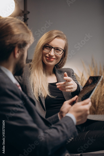 Charming woman with blond hair sitting on grey couch with bearded man that holding digital tablet. Two business partners in formal outfit having conversation at office.