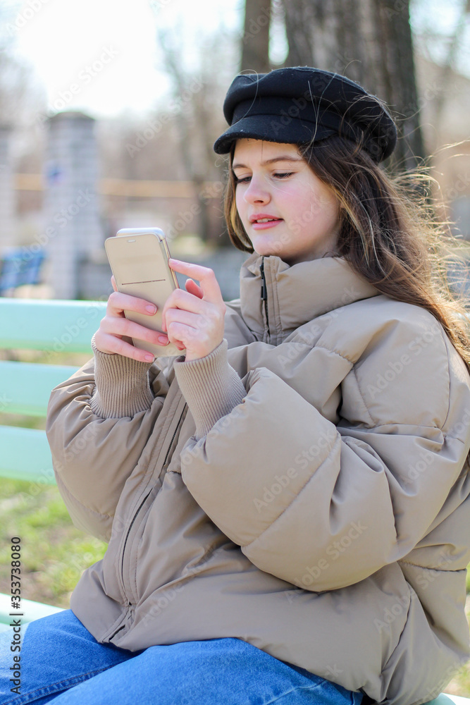 Long-haired girl in a cap holds a smartphone while sitting on a bench in the park Long-haired girl in a cap holds a smartphone while sitting on a bench in the park