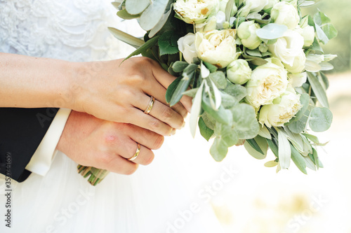 Closeup view of newlyweds hands holding colorful wedding bouquet. Bride and groom wearing wedding rings. Outdoor background. Wedding day concept.