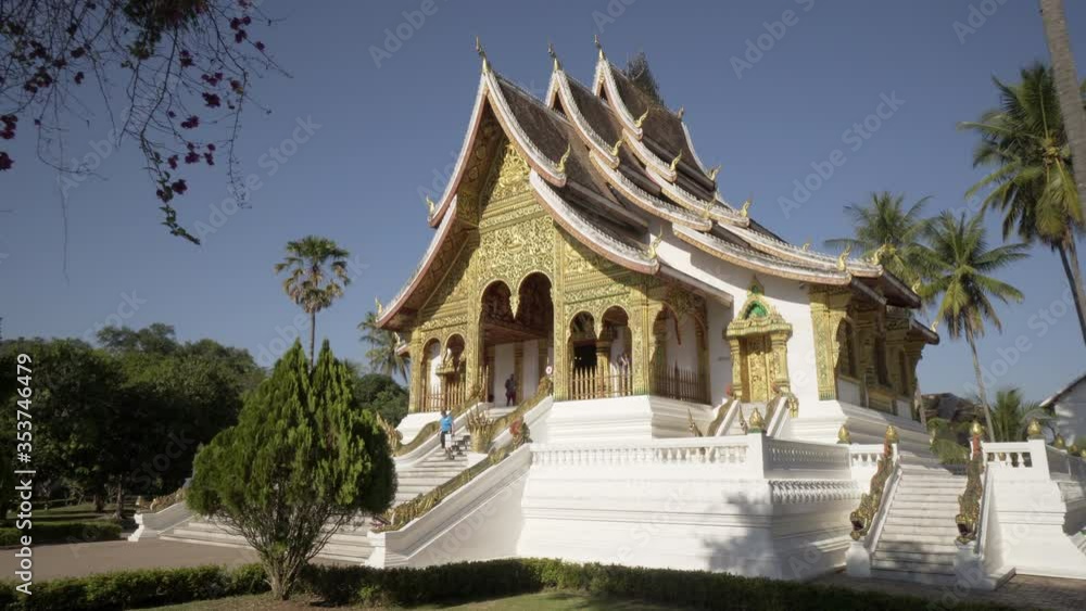 Panning shot of famous temple with tourists amidst palm trees against sky during sunny day - Luang Phabang, Laos Panning shot of famous temple with tourists amidst palm trees against sky during sunny day - Luang Phabang, Laos