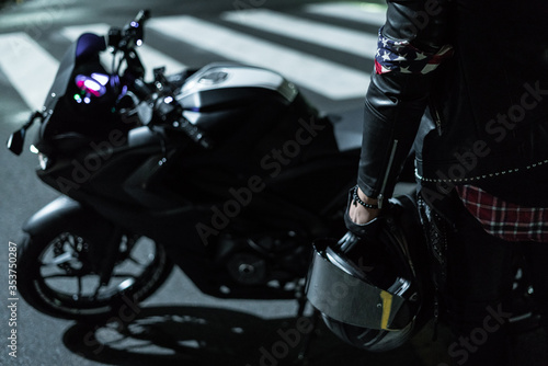 Piloto de moto con su moto detenido en la noche en la calle de la ciudad con una pañoleta en su brazo izquierdo sosteniendo su casco y su moto en el fondo de la toma
