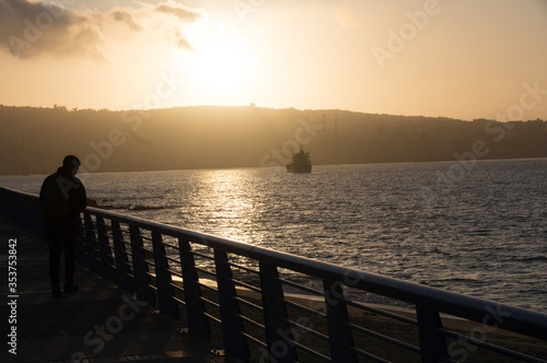 Mar desde la costa en un muelle en un atardecer donde se pueden ver los barcos y un cerro en el fondo donde se oculta el sol