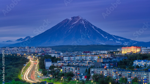 Panorama of night city of Petropavlovsk-Kamchatsky on background cone of volcano, urban development at twilight, backlit city road with driving car lights.