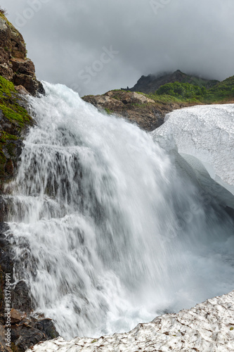 Scenery view of idyllic cascade waterfall falling into snowfield in rocky mount