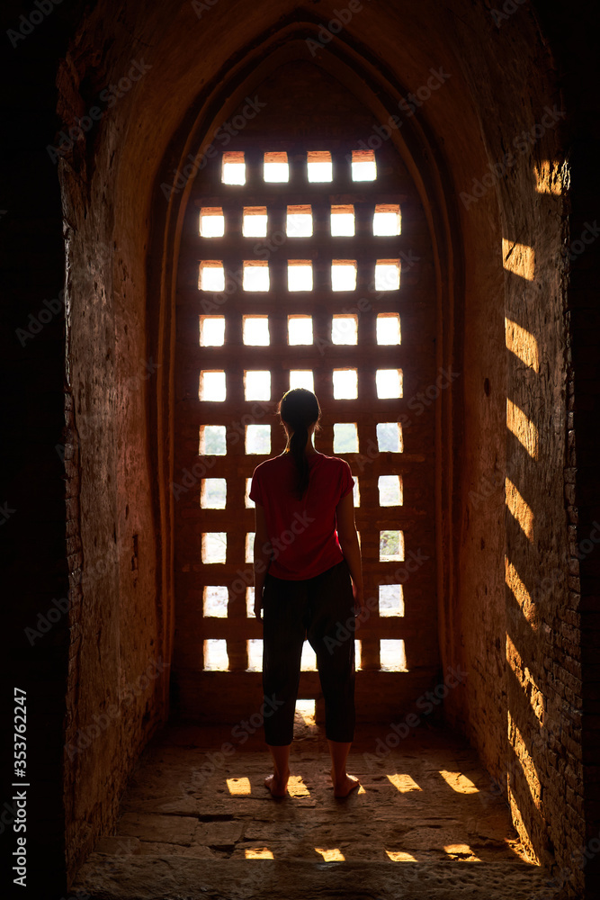 Sunset light falls on a girl traveler through a lattice window in an ancient temple in Bagan, Myanmar, Burma.