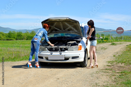 Red-haired and brunette women on high heels with a screwdriver and pliers, standing near a broken car with opened hood on a country road and barrier with stop sign