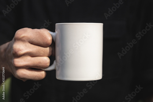Man holding a white cup of hot beverage outdoors. Man's hand holding a blank mug against black background. Close up, mockup image, copy space for text.