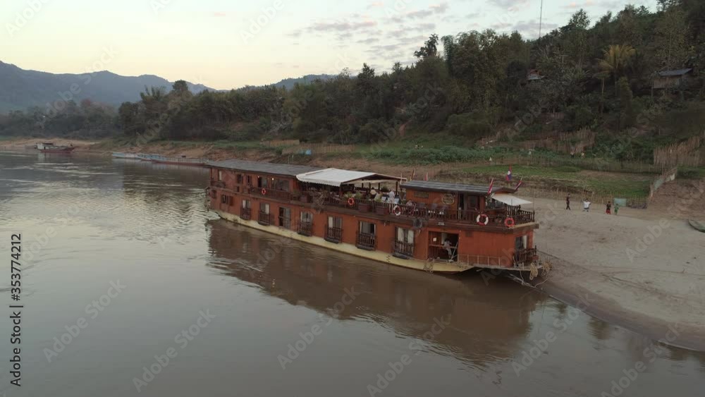 Aerial shot of cruise ship at riverbank by people near trees, drone ascending over river against sky during sunset - Mekong River, Laos Aerial shot of cruise ship at riverbank by people near trees, drone ascending over river against sky during sunset - Mekong River, Laos