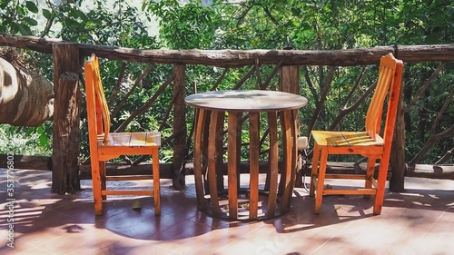 Wooden table and two wooden chairs Laid bare, no one sitting Arranged for sitting, relaxing or eating The area near the wooden fence in front of the house With trees and leaves Object background