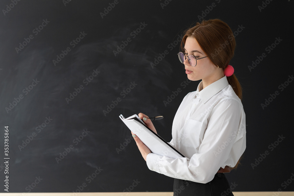Young beautiful woman teacher stands near a school blackboard. Writing in the notebook all the most important. Free space for text. Young beautiful woman teacher stands near a school blackboard. Writing in the notebook all the most important. Free space for text.