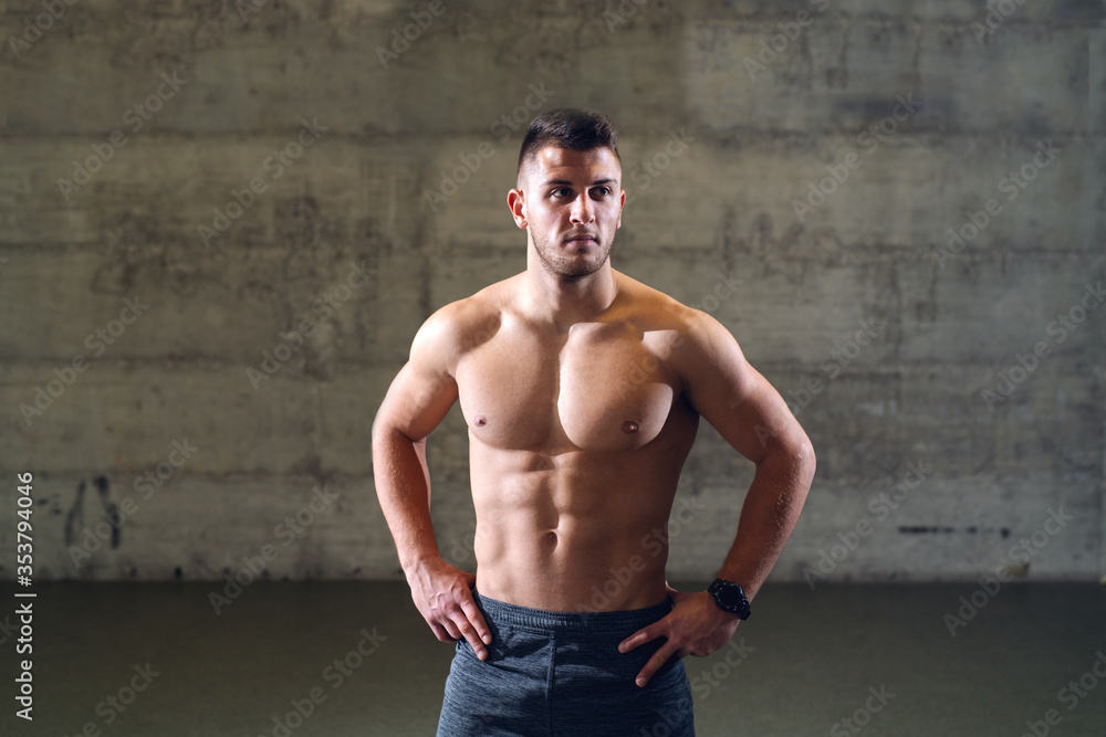 Shirtless serious Caucasian man with hands on hips posing while standing in front of the wall.