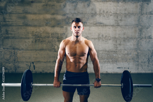 Serious Caucasian shirtless man in shorts lifting barbell. In background wall.