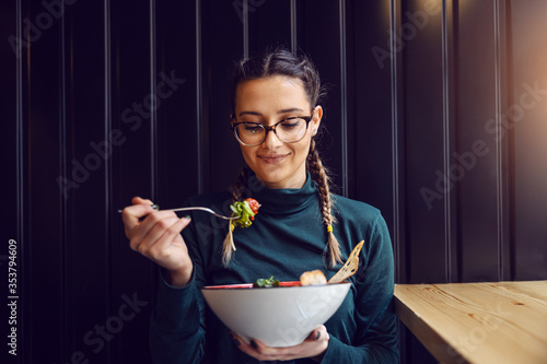 Young attractive teenage girl with healthy habits sitting in restaurant, holding bowl with salad and having lunch. photo