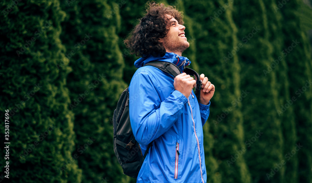 Side view image of the cheerful young man with curly hair carrying backpack looking up to the sky. Attractive male resting in the street on a rainy day. Hipster guy has joyful emotion. Side view image of the cheerful young man with curly hair carrying backpack looking up to the sky. Attractive male resting in the street on a rainy day. Hipster guy has joyful emotion.