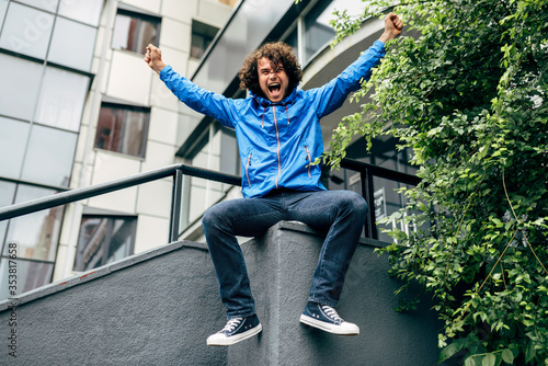 Outdoor image of the cheerful young man with curly hair sitting on stairs of a building with raised hands up. Attractive male resting in the street on a rainy day. Hipster guy has joyful emotion.