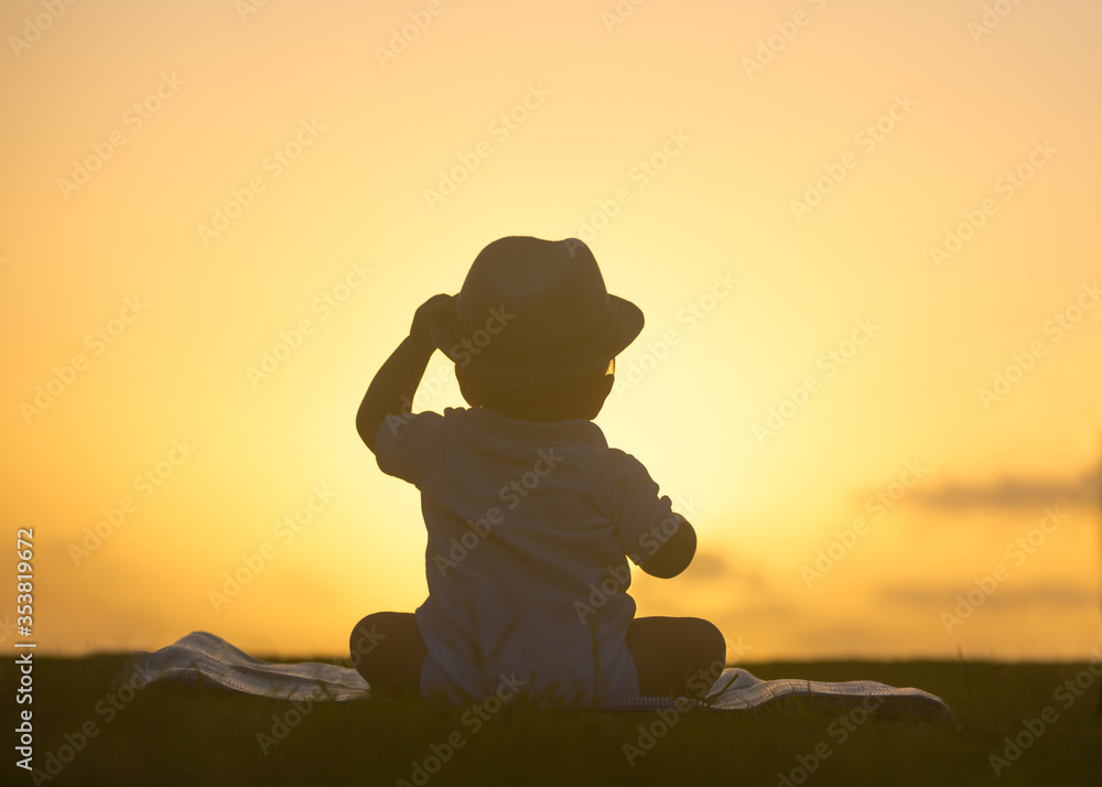 Silhouette portrait of cute baby boy wearing a gentleman's hat while sitting on the grass at the park during beautiful yellow color sunset.