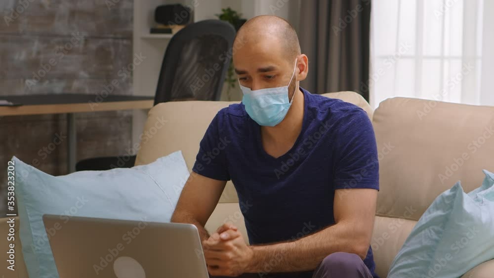 Man with protection mask waving during a video call on laptop. Coronavirus self isolation.